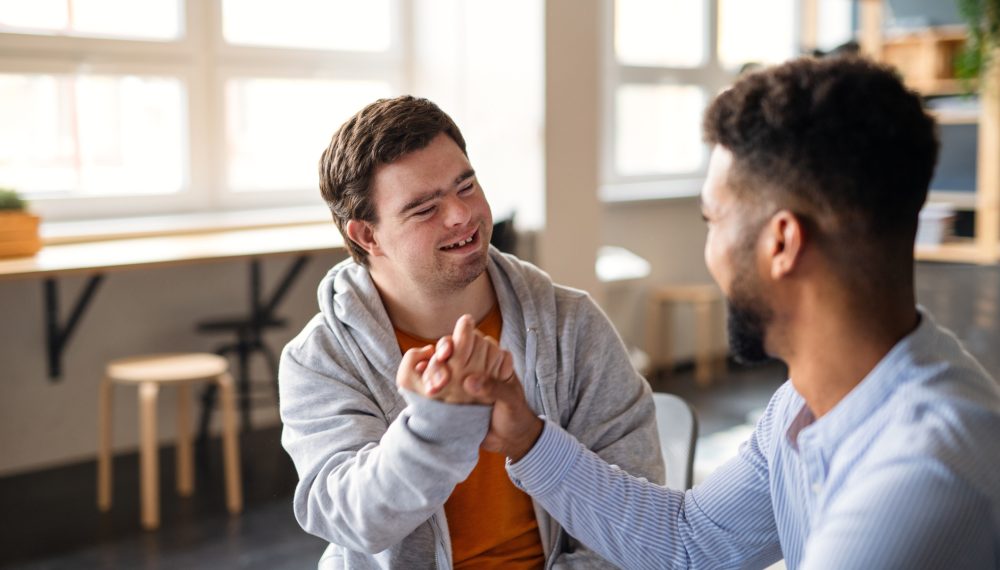 A young happy man with Down syndrome with his mentoring friend celebrating success indoors at school.